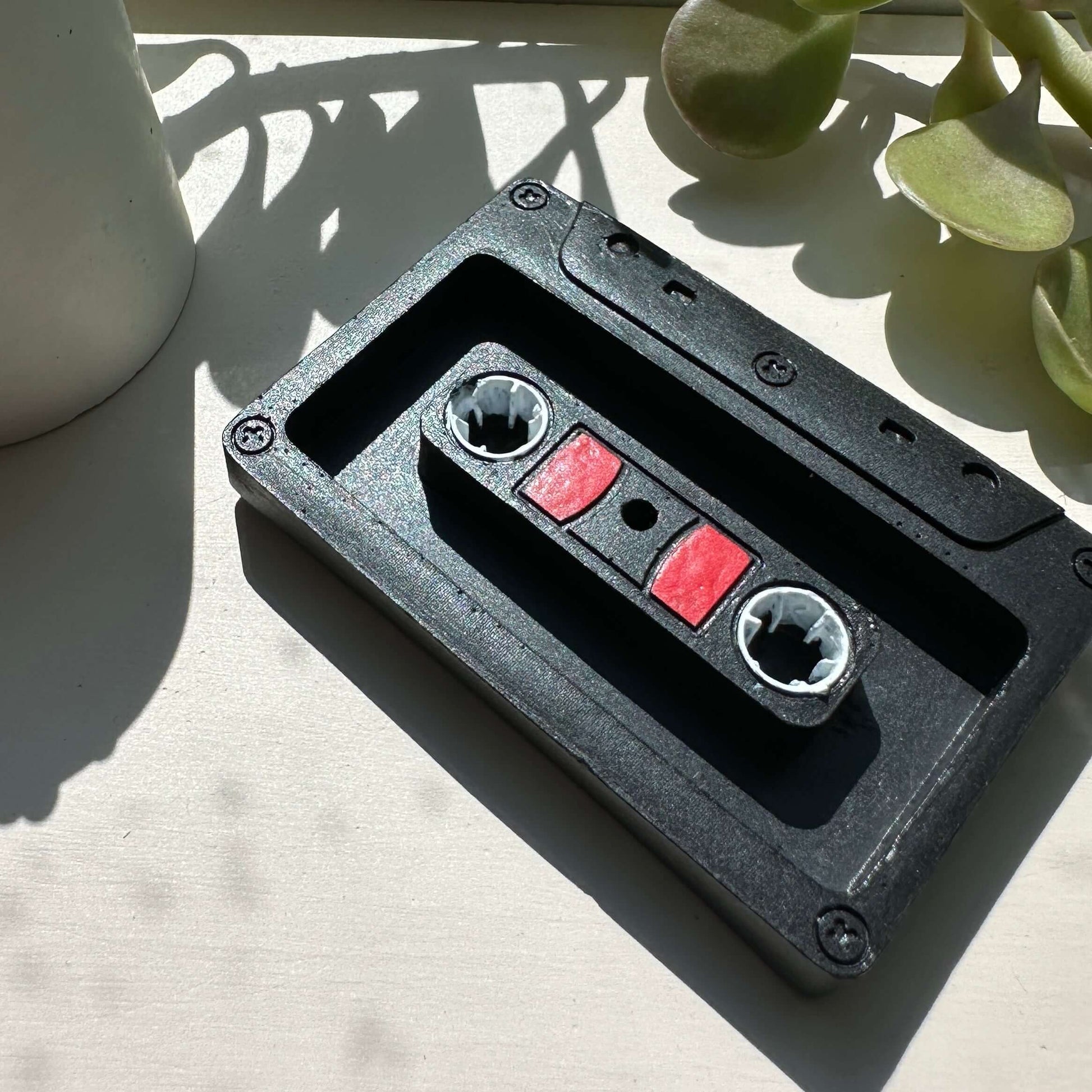 Ceramic cassette tape incense stick holder on a white table next to green succulent plants.