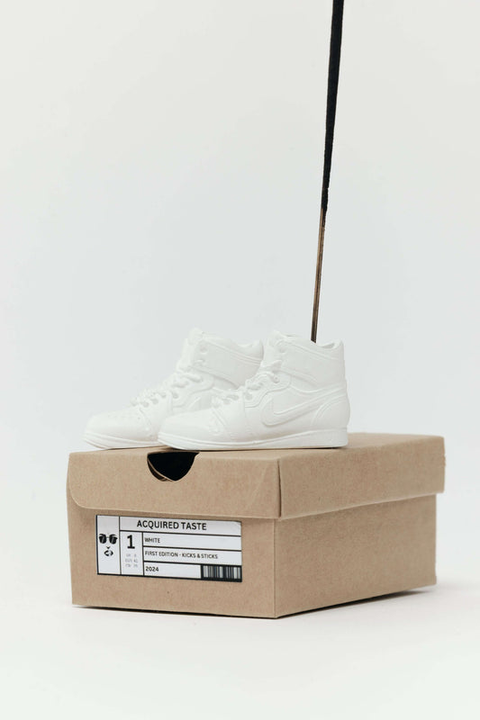 White sneakers on a cardboard box with 'Acquired Taste' label against a white background ncense holder